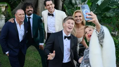 Group of six people in formal attire taking a cheerful outdoor selfie with greenery in the background.