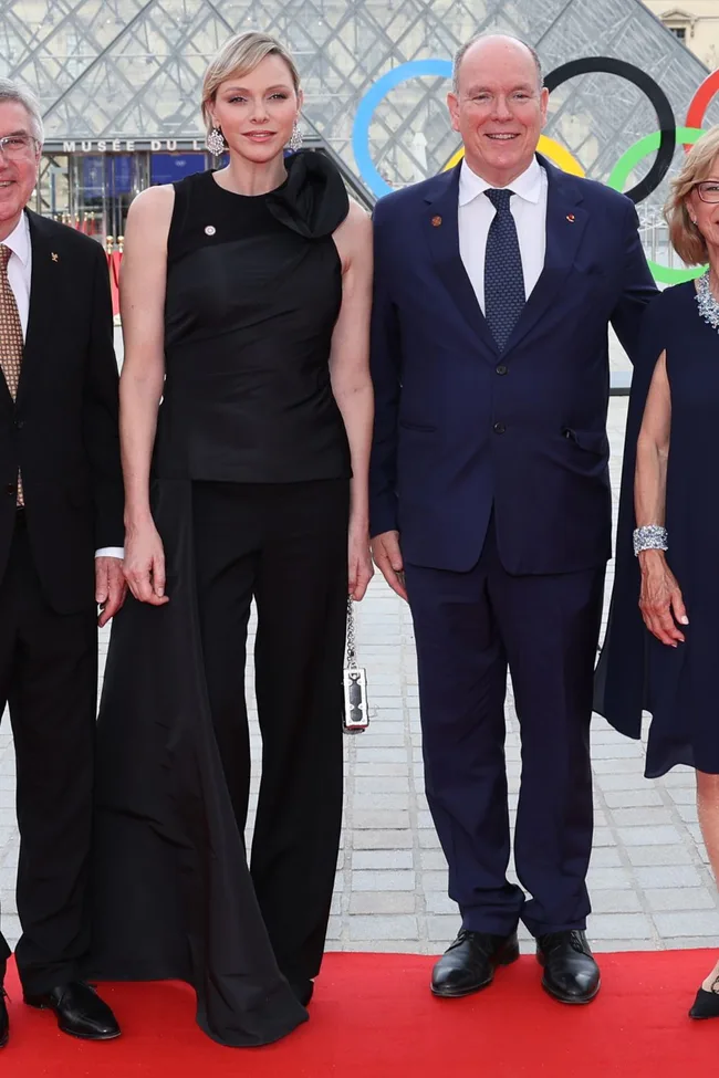 "Group on red carpet outside Louvre, Paris. Elegant attire, Olympic rings visible in background."