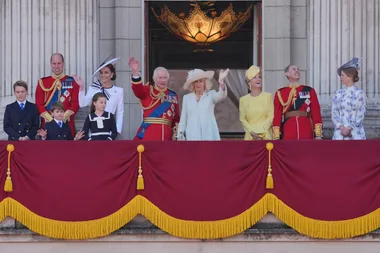 A royal family on a balcony waving, dressed in formal attire, with a red and gold decorated backdrop.