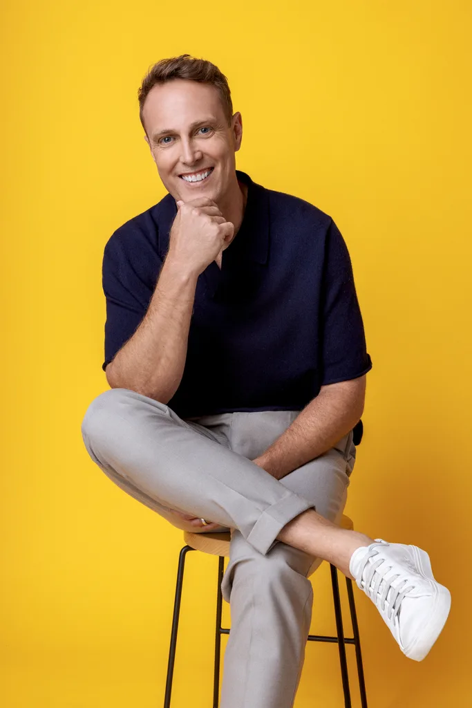 Man seated on stool against a yellow background, smiling, wearing a navy shirt, gray pants, and white sneakers.