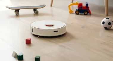 Robot vacuum cleans wooden floor surrounded by kids' toys, including a skateboard and a small soccer ball.