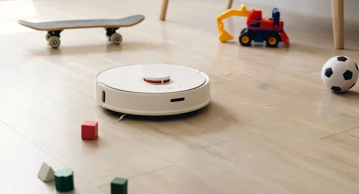 Robot vacuum cleans wooden floor surrounded by kids' toys, including a skateboard and a small soccer ball.