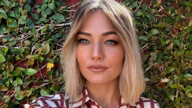 sam frost with blonde hair and a red and white blouse standing in front of a hedge