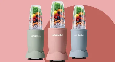 Three Nutribullet blenders in pastel colors, filled with fruits, nuts, and greens on a pink background.