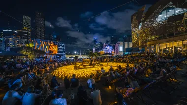 Outdoor candlelight concert at night, with a crowd seated around, surrounded by city skyscrapers.