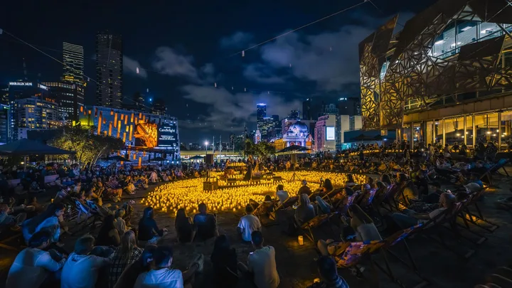 Outdoor candlelight concert at night, with a crowd seated around, surrounded by city skyscrapers.