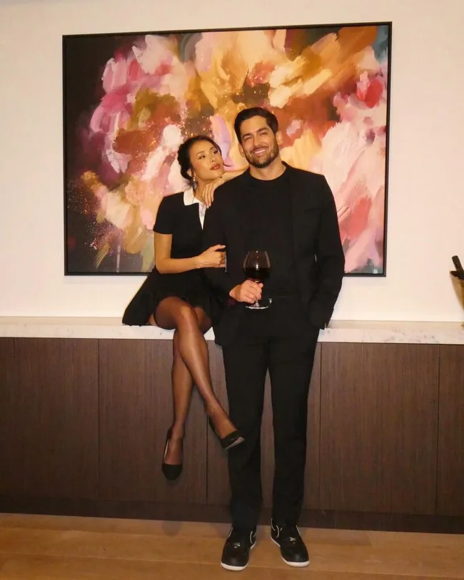 A couple dressed in black, with one seated on a counter, in front of a colorful abstract painting.