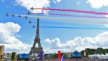 Seven jets fly over the Eiffel Tower, leaving red, white, and blue trails, with a crowd below on a sunny day.