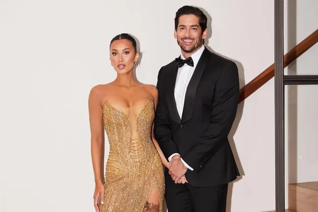 A couple posing elegantly; woman in gold dress, man in black tuxedo, both smiling.