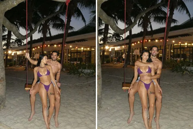 A couple posing on a swing at sunset on a beach with palm trees and festive lights in the background.