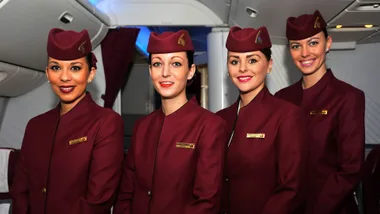 Four flight attendants in burgundy uniforms and hats standing inside an airplane cabin, smiling.