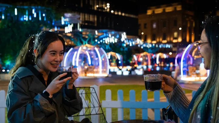 Two people enjoying hot drinks at an outdoor event with colorful illuminated domes in the background at night.