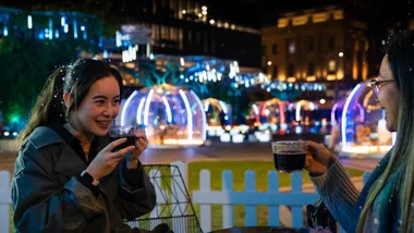 Two people enjoying hot drinks at an outdoor event with colorful illuminated domes in the background at night.