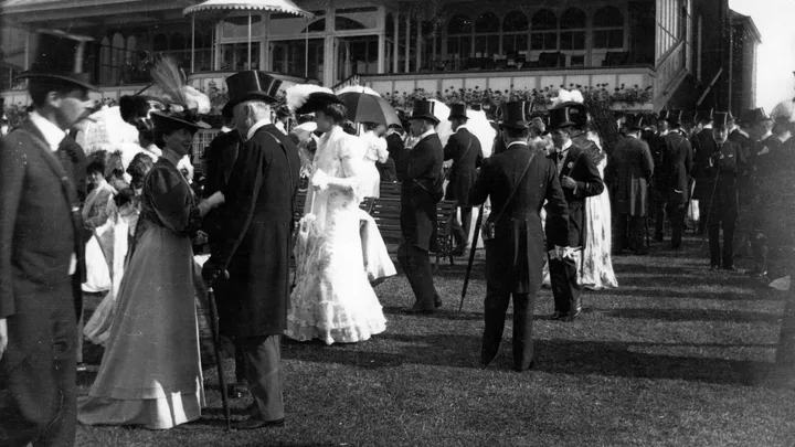 Black and white photo of elegantly dressed people in Edwardian fashion, socializing outdoors with a large building in background.