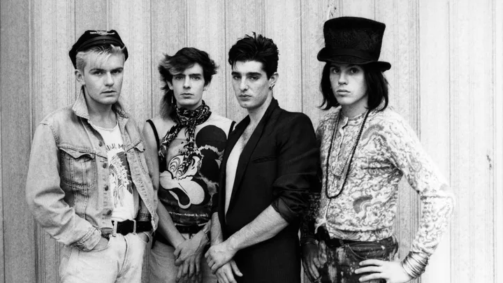 Black and white photo of four men in '80s fashion, standing against a striped wall, one wearing a tall top hat.