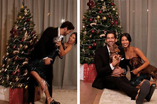 A couple dressed elegantly by a Christmas tree; smiling, holding wine and dogs, creating a festive atmosphere.