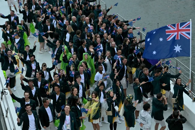 Australian team with flags and uniforms at an outdoor event, waving and smiling on a white platform by water.
