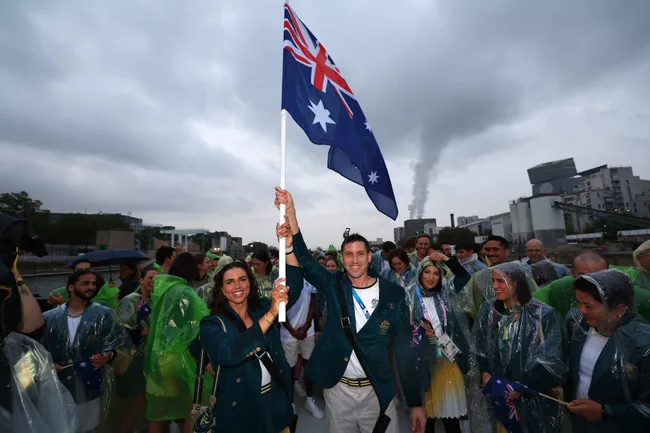 Australian team holding their national flag at an outdoor event, wearing green and gold with raincoats.