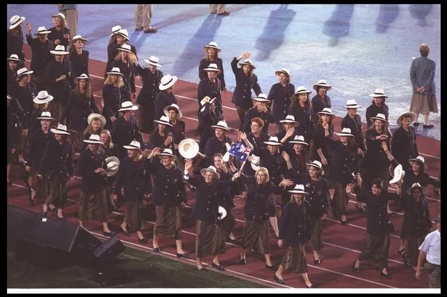 Australian athletes in formal attire, waving and carrying flags, march on a stadium track during an opening ceremony.