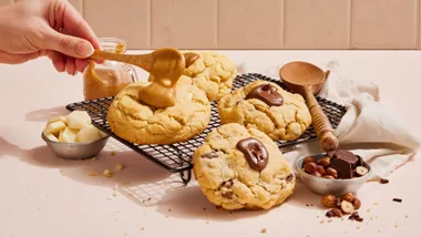 Hand spreading caramel over cookies on a cooling rack, surrounded by chocolate and nuts.
