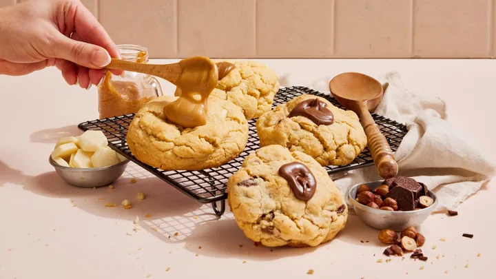 Hand spreading caramel over cookies on a cooling rack, surrounded by chocolate and nuts.