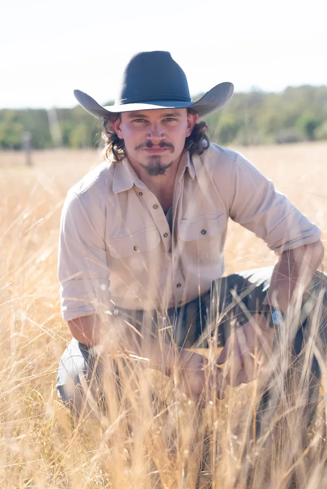 Man in cowboy hat and shirt kneels in a dry grassy field, sunlight casting shadows on his face.