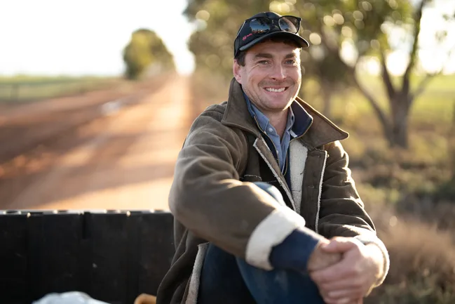 Man in jacket and cap smiling, seated on a rural road with trees and blurred background.
