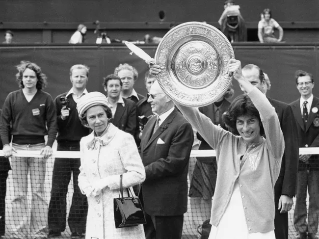 A woman holds a trophy aloft at a tennis event, with an elegantly dressed lady beside her and a crowd in the background.