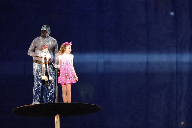 A man covered in white powder stands on a platform holding a stick while a girl in a pink dress with a hair bow stands beside him.