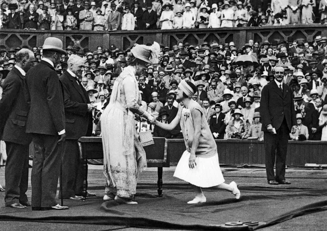 A woman curtsies and shakes hands with another woman in front of a crowd at a historic tennis event.