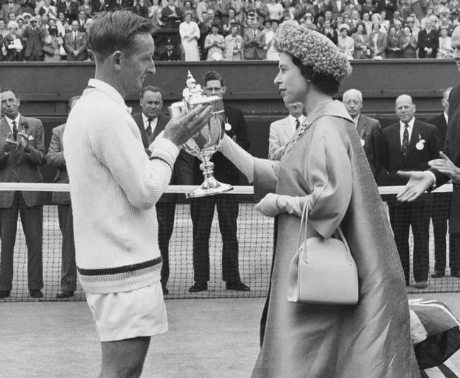A man in tennis attire receives a trophy from a woman wearing a cape and hat on a tennis court, with a crowd watching.