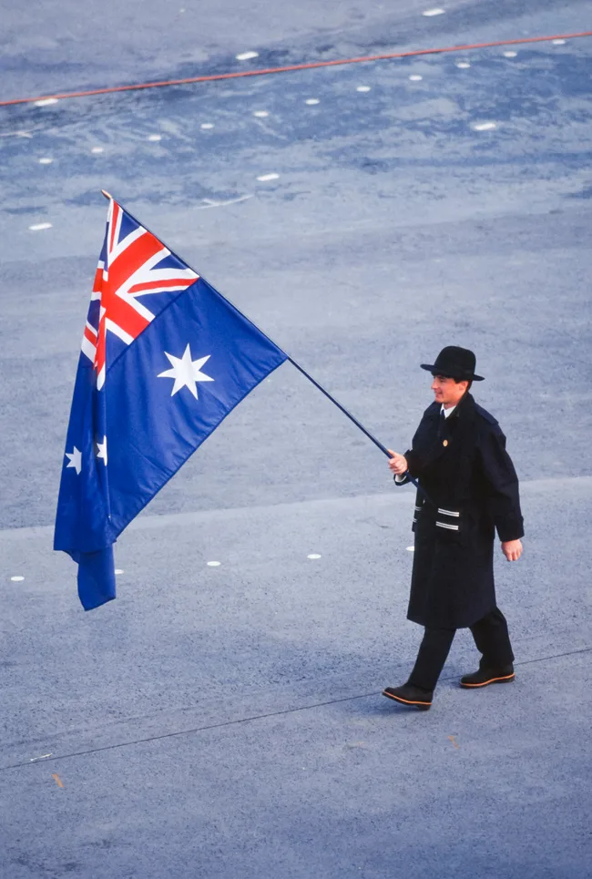 Man in a dark coat and hat walking on a tarmac carrying the Australian flag.