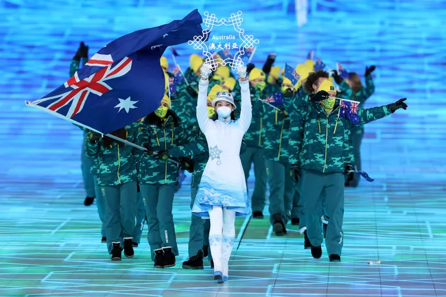 Australian Olympic team entering ceremony, flag bearer leading in white outfit, blue flag with stars, winter sports theme.
