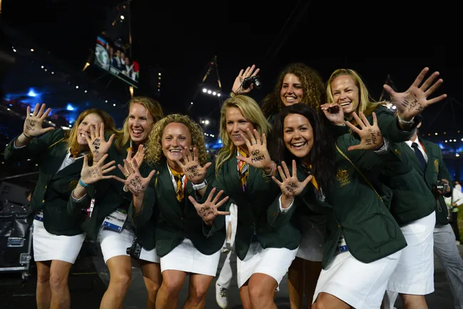 Australian Olympic team members smiling, wearing green blazers and white skirts, showing messages written on their palms.