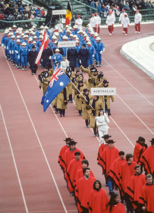 Participants in colorful outfits and flags march during an Olympic-style opening ceremony on a track.