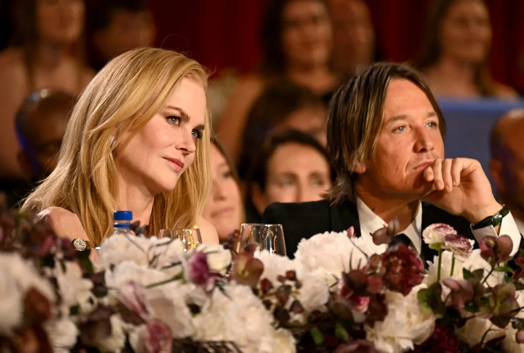 Two individuals seated at a table with floral decor, attentively watching an event in a formal setting.