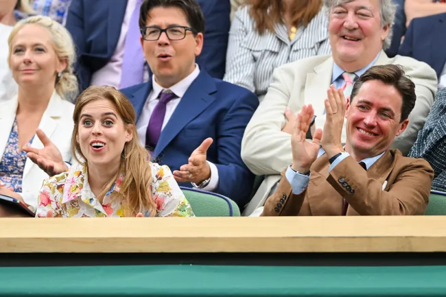 Group of people in a stadium, some clapping and others reacting with surprise and smiles.
