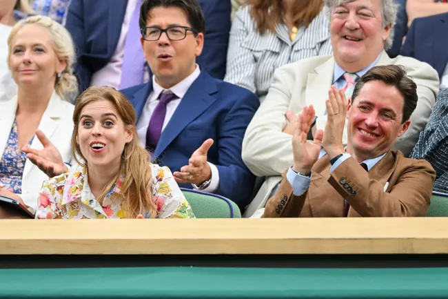Group of people in a stadium, some clapping and others reacting with surprise and smiles.