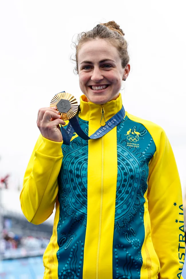 Australian athlete smiling, holding a silver medal, wearing a yellow and blue jacket with "PARIS 2024" lanyard.