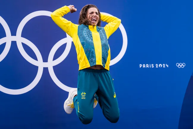 Athlete in yellow and green jumps joyfully in front of Paris 2024 Olympic rings backdrop.