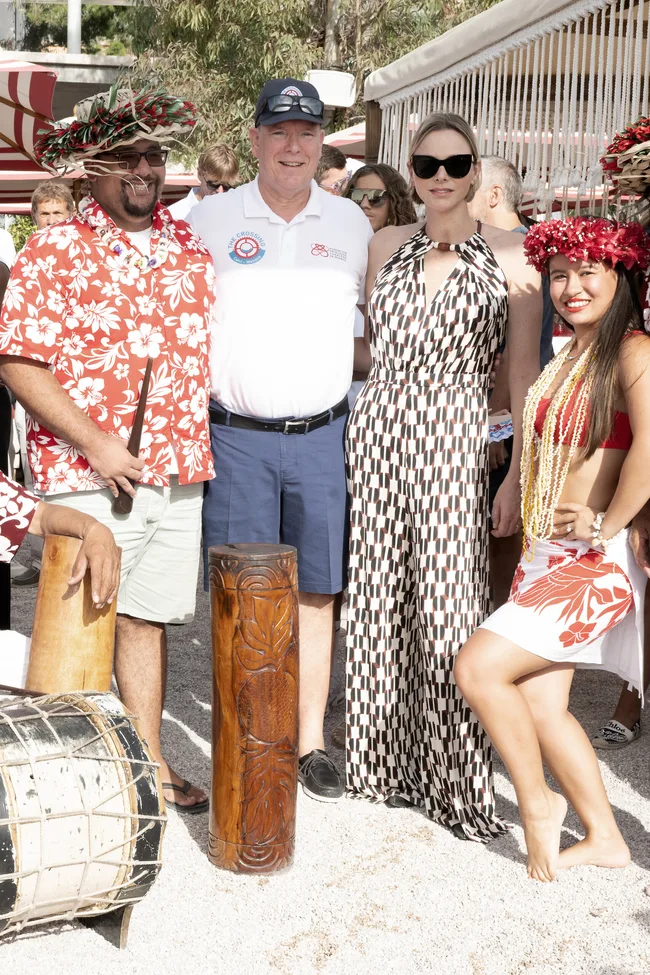 A group of people standing together in summer attire, one in a printed dress and sunglasses, surrounded by traditional elements.