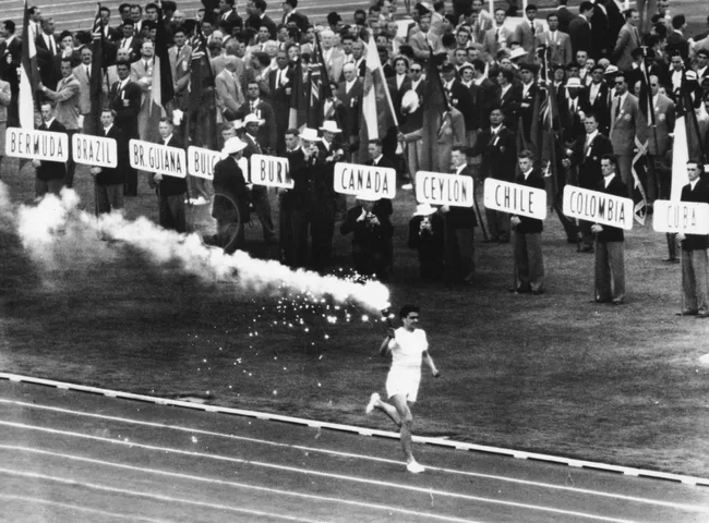 Athlete running with a lit torch at the Olympic opening ceremony, with officials and flags in the background.
