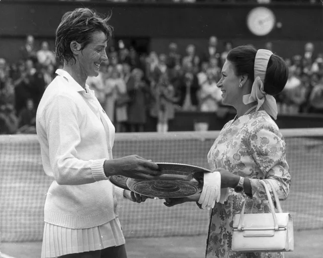 Tennis player accepts trophy from a woman at a court ceremony, with a crowd in the background applauding.