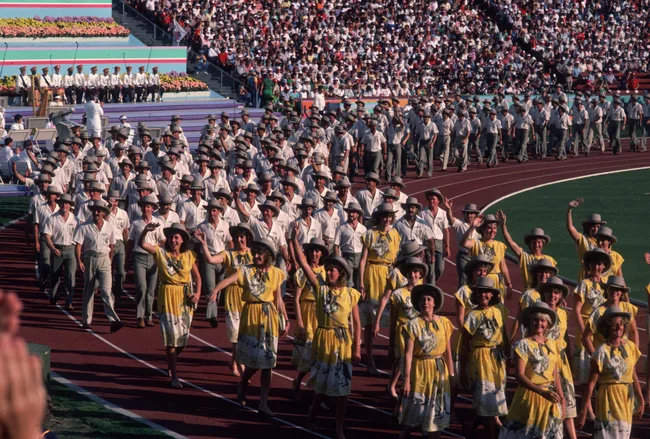 Crowd of athletes in white and yellow outfits and hats marching on a stadium track during an event.