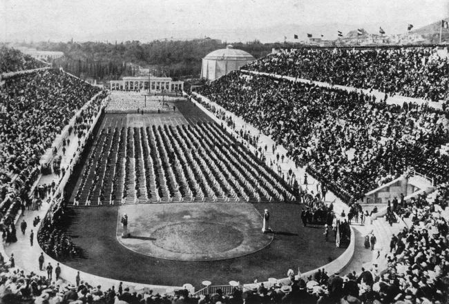 Crowd at the 1896 Summer Olympics in Athens, Greece, seated in a stadium with athletes on the field.