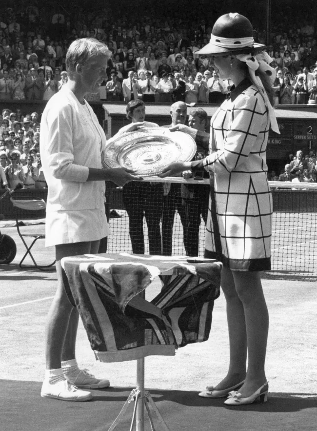 Tennis player receives trophy from a woman in a hat at a crowded tennis court trophy ceremony.