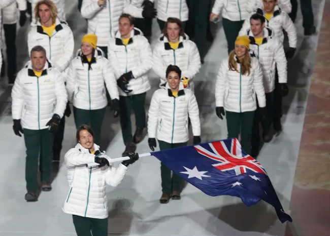 Australian Olympic team walking in a ceremony, wearing white jackets, one person holding an Australian flag.