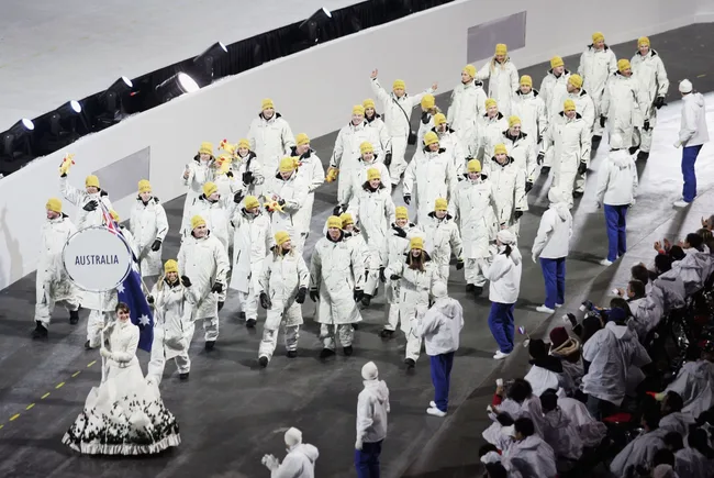 Australian team in white winter outfits and yellow hats march at a ceremony, led by a flag-bearer with an Australia sign.