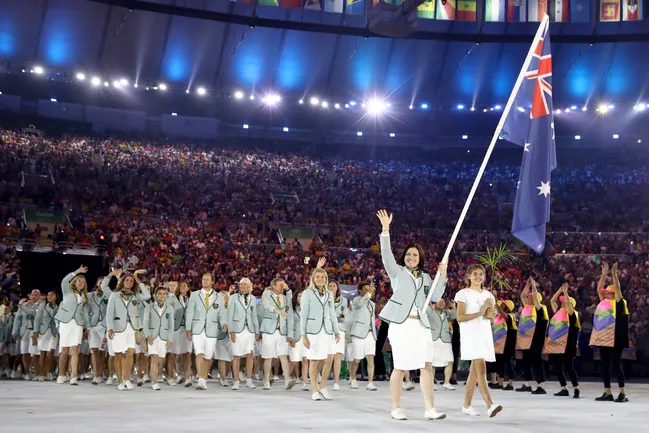 Australian athletes in uniforms parade at the Olympics opening ceremony, holding the national flag, with a large crowd watching.