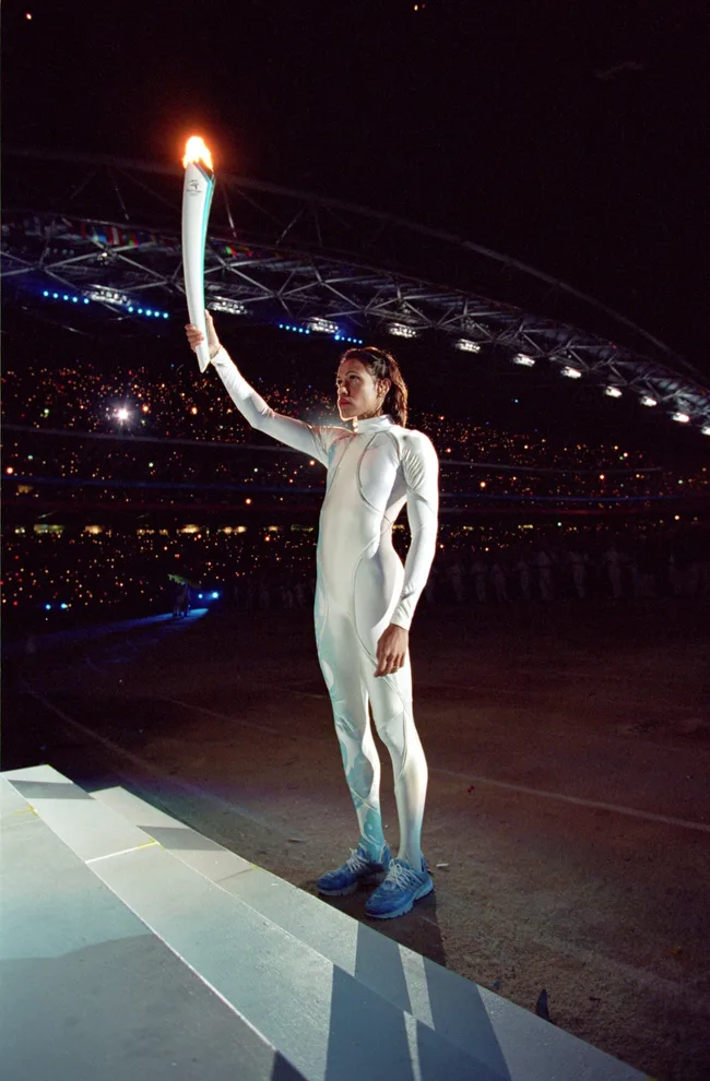 Olympic athlete holding a flaming torch in a stadium at night, standing on steps in a fitted white outfit and blue shoes.